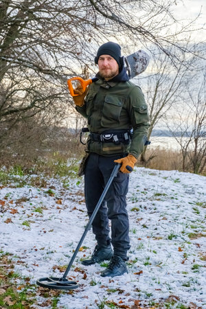 Full body young caucasian bearded man with a metal detector and a shovel, looking for treasure in snowy forest in winter. Vertical orientationの写真素材