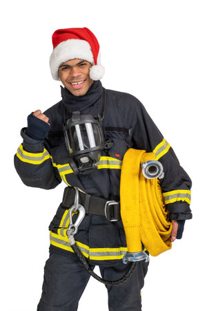 Exited young African American fireman in uniform and red Santa hat holding fire hose and looking at camera isolated on white backgroundの写真素材