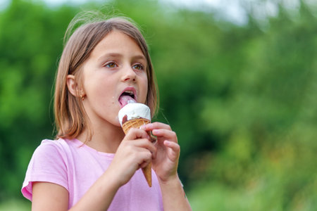 Cute little girl eating an ice cream cone holding it with both hands in summer in the park. High quality photo, blurred backgroundの写真素材