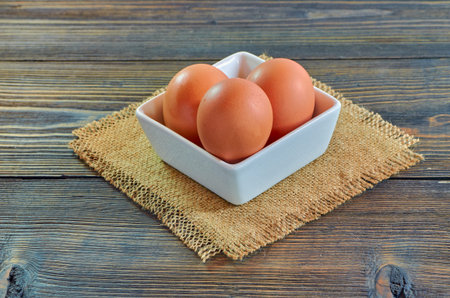 Chicken eggs in a square white clay plate on a burlap napkin, on a wooden background.の写真素材
