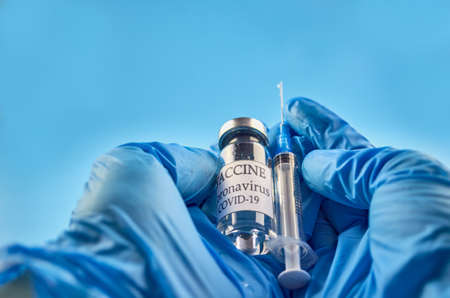 A bottle of COVID-19 vaccine and a syringe with an injection needle against coronavirus infection in the doctor's hand in a nitrile glove on a blue background. Close-up. Place to copy. The concept of medicine, health care, vaccination and immunization. High quality photoの写真素材