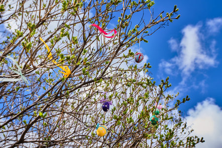 Easter multi-colored eggs hang from a tree in the garden against a blue sky. Sunny spring day. The...