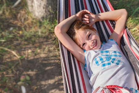 A happy little girl lies in a hammock in the garden in the summer. A child in a hammock during the holidays smiles in the park against the background of green grass. High quality photoの写真素材