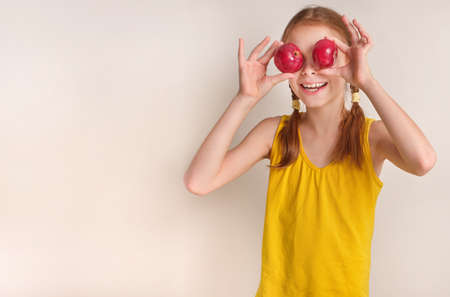 A positive playful girl with apples on a gray-white background. A happy child, a smiling girl holding fresh red ripe apples in her hand. There is a basket of apples nearby. The concept of healthy and organic nutrition.の写真素材