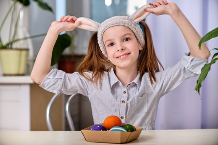 A positive playful girl in bunny ears holds colorful eggs in her hands in the home kitchen. Happy Easter. Easter backgroundの写真素材