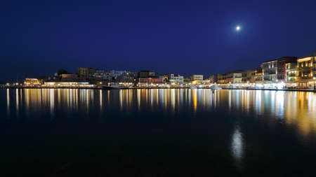 Night skyline of Chania town, Crete, sea bay, buildings beautifully illuminated, full moon and starry sky, reflections in waterの写真素材