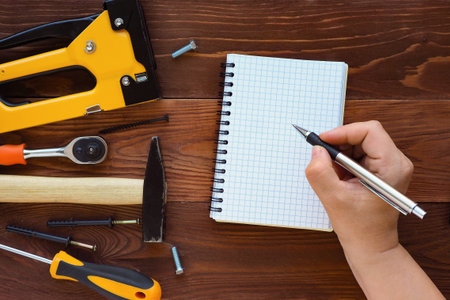 hand with pen writes in a notebook on a dark wooden background and tools for repair and...