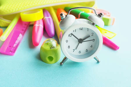 School office supplies: felt-tip pens, markers, sharpener, eraser, pens, tape, rulers of bright neon colors drop out of a yellow pencil case on a blue paper background and a white alarm clock.の写真素材
