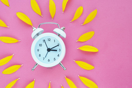A white alarm clock on pink colored paper and many yellow petals around. space for text.