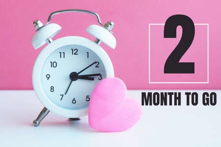 A small white alarm clock and a pink heart figurine. The inscription "2 month to go".の写真素材