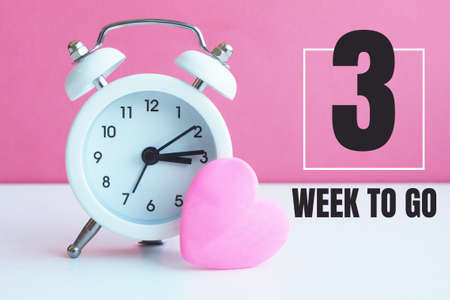 A small white alarm clock and a pink heart figurine. The inscription "3 week to go".の写真素材