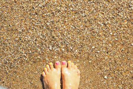 Female legs with bright pink and yellow nails on the sand in the sea water.の写真素材