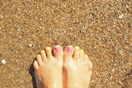 Female legs with bright pink and yellow nails on the sand in the sea water.