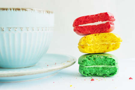 Three bitten macaroon cakes of red yellow and green colors in a stack near a blue cup on a white table.の写真素材