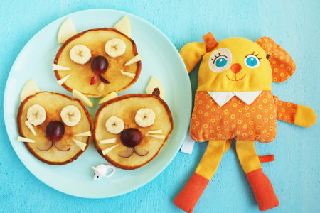 A plate with three pancakes in the shape of cat faces, decorated with slices of banana, grape and apple. orange toy dog. Children's food serving. Art food.の写真素材