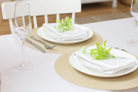 Plates with twigs of artificial flowers on beige round napkins, glasses and cutlery on a white...