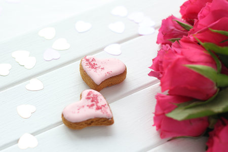 two gingerbread hearts on a wooden background, confetti hearts and a bouquet of bright pink roses.