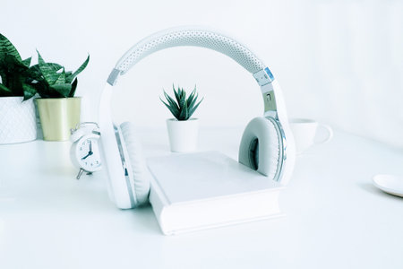 white wireless headphones on a white paper, houseplants, a cup and a small white alarm clock on a...