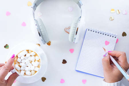 female hands with beautiful manicure hold a cup of coffee and marshmallows and a pen. White headphones with a notepad on a white background with confetti hearts. top view, lifestyle.の写真素材