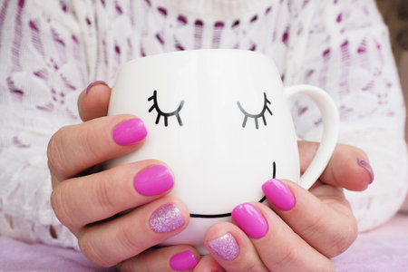 female hands with beautiful lilac manicure hold a white mug with a painted muzzle.