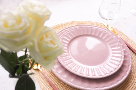 table setting. two empty pink plates ,, roses in a vase, spoon and fork on a white table. copy...