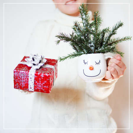 a girl in a white sweater holds out a white mug with a snowman's face and fir branches and a red gift box.の写真素材