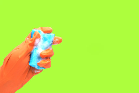 a hand in an orange rubber glove and a blue sponge with foam on a green background.