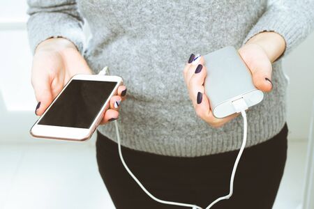 Two girls hands hold of the phone and charger. Powerbank and smartphone in girl's hands. Energy charger power bank smart phone.の写真素材