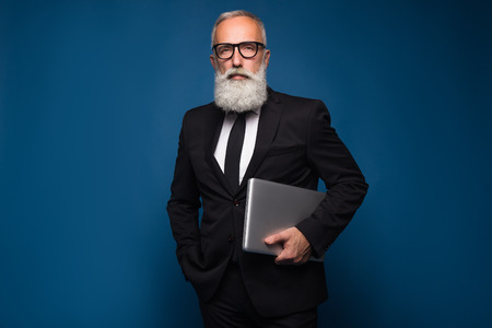 Concentrate bearded senior and business man staying with notebook and working isolated on a blue background. Working business man in straight suit and glassesの写真素材