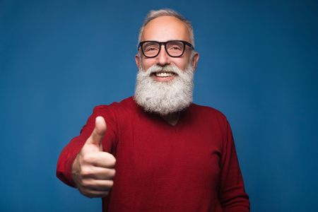 Happy modern old bearded man in warm sweeter show thumb up and happy smiling. Isolate on blue background. Successful and handsome adult bearded man of enjoying him emotions. Show ok and happy emotionsの写真素材