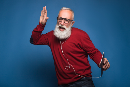 Expectation from a new song. Happy modern bearded man listening a new music from tablet. Man with headphones listen to music isolated on blue background. Happily dances from cool music on headphones.の写真素材