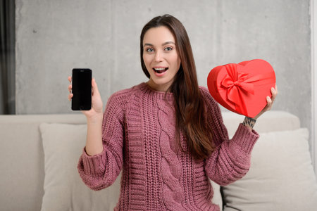 portrait of a beautiful girl wearing sweater and holding heart-shaped gift box and mobile phone