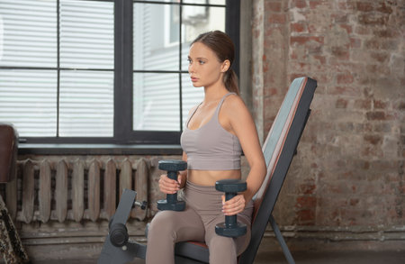 Fit athletic woman in leggings and top sitting on exercise machine, holding dumbbells, training and looking ahead on a window and brick wall background. sport concept.の写真素材