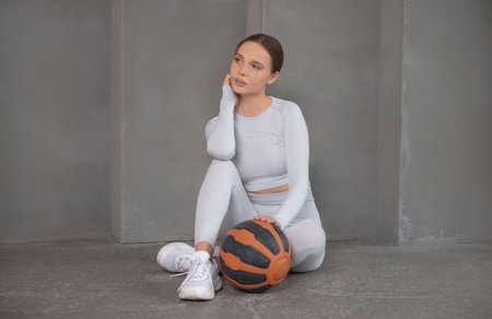 Young fit woman wearing white sportswear sitting on floor, holding basketball ball and looking aside on a gray background. Sporty woman taking break after workout.の写真素材