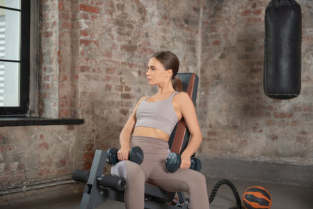 Young fit athletic woman in leggings and top sitting on exercise machine, holding dumbbells, training and looking ahead on a window, punching bag and brick wall background.の写真素材