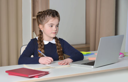 Cute girl in school uniform having video conference with teacher from home using laptop and writing in notebook. School kid looking at computer screen and taking notes.の写真素材
