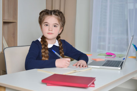 Cute school girl with pigtails and school uniform looking at camera, making notes or writing in...