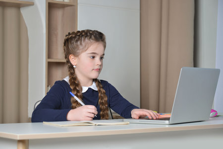 Cute school girl having online lesson using laptop and keyboard, looking at computer screen and taking notes or writing in exercise book. Back to school and learning concept.の写真素材