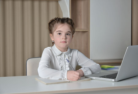 Cute schoolgirl taking notes or writing in notebook, looking at camera and sitting at desk on a laptop and multicolored paper clips stickers background. Back to school concept.の写真素材