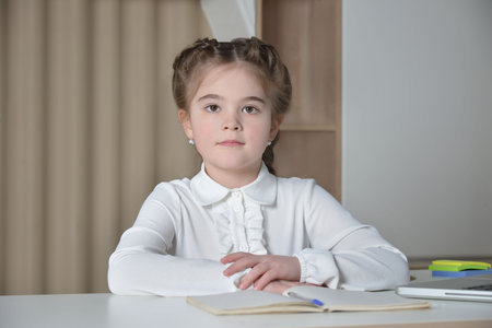 Cute little girl from elementary school sitting at desk and looking at camera on a notebook, pen, paper clips stickers and curtains background. Back to school concept.の写真素材