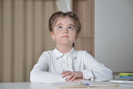 Cute schoolgirl in white shirt looking up and sitting at desk. Portrait of caucasian young girl from elementary or primary school. Homeschooling and back to school concept.の写真素材