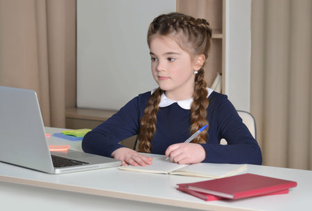 Cute girl in school uniform having video conference using laptop and writing in notebook at home. School kid looking at computer screen and taking notes. Online education concept.の写真素材