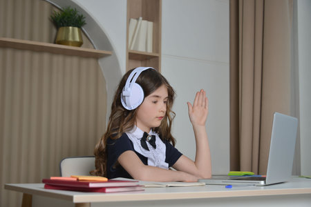 Schoolgirl raising hand wearing wireless headset or headphones, studying from home in front of laptop and sitting at table. Secondary student having online lesson at home.の写真素材