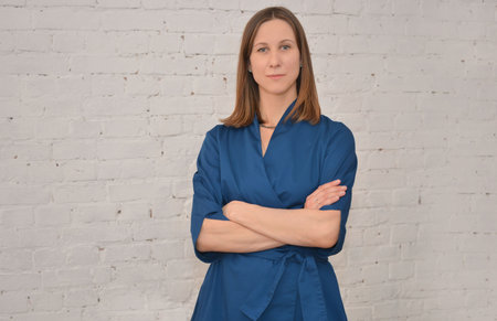 Middle aged female doctor wearing blue uniform, looking at camera and holding hands crossed on chest on a white wall background. Medical service, medicine or treatment concept.の写真素材