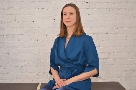 Middle aged or mature female doctor wearing blue uniform, looking at camera and holding hands on knees on a white brick wall background. Medical service, medicine or therapy.の写真素材