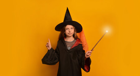 Child girl in wizard costume with luminous magic wand looking at camera and pointing finger up on yellow background.の写真素材