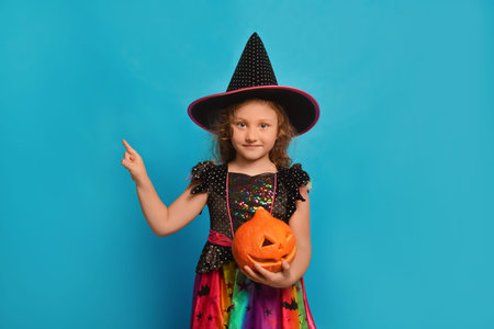 Little girl in witch costume with Halloween pumpkin pointing index finger up or holding finger aside on blue background.の写真素材