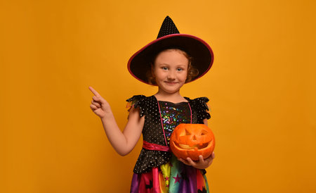 Cute kid girl in witch costume holding pumpkin, pointing finger aside and looking at camera on yellow background.の写真素材