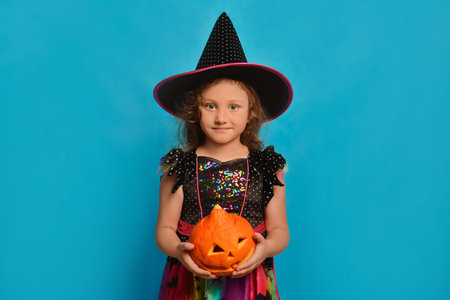 Little witch girl holding Halloween pumpkin in hands and looking at camera on a blue background.の写真素材