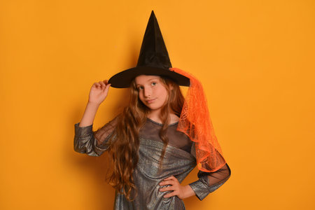 Teen girl in Halloween witch costume holding hat, keeping hands on waist and looking at camera on yellow background.の写真素材